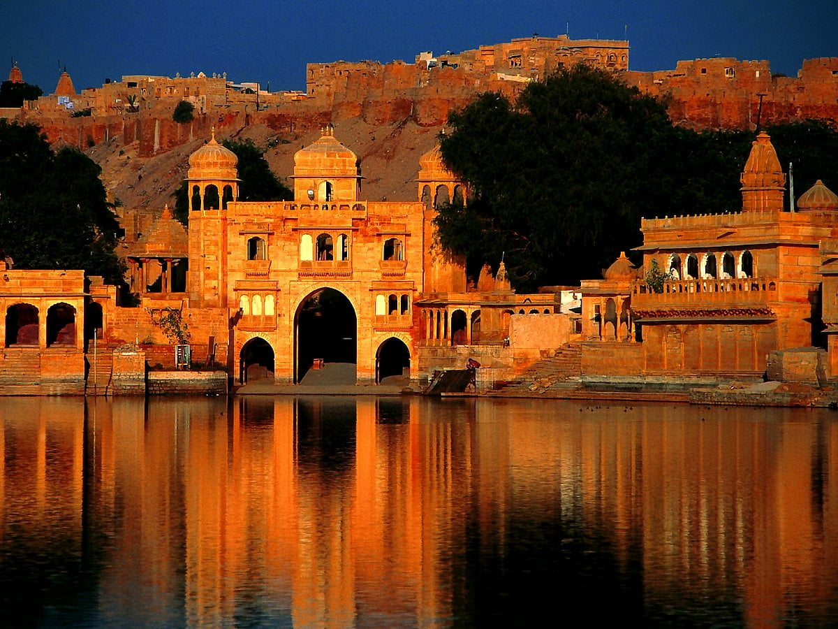 Yellow sandstone temple gateways and arches beside a lake, with a hilltop fort visible behind at dusk