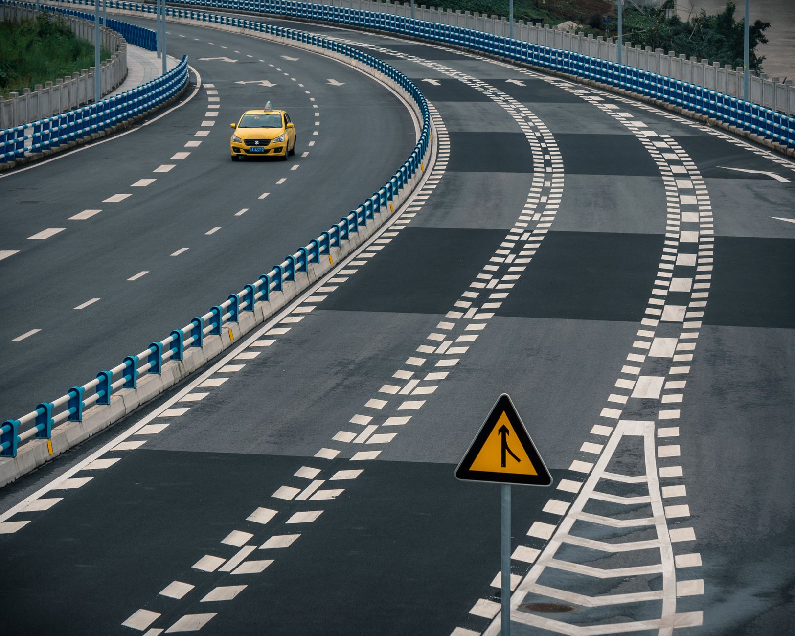 A lone yellow taxi navigates around a curved, empty highway with a merge sign visible.