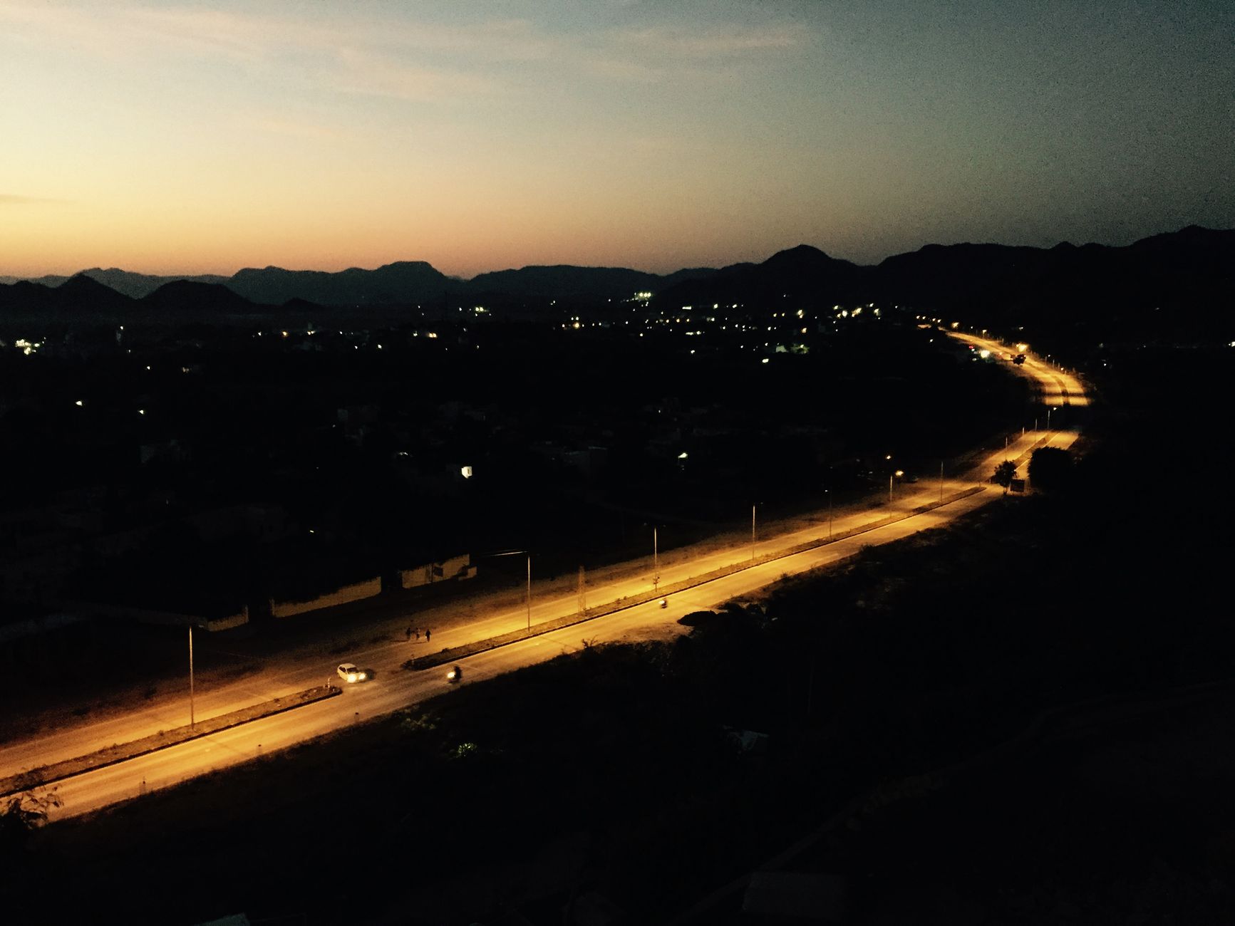 A serene night view of a lit highway in Sukher, Rajasthan, India, with mountains in the backdrop.